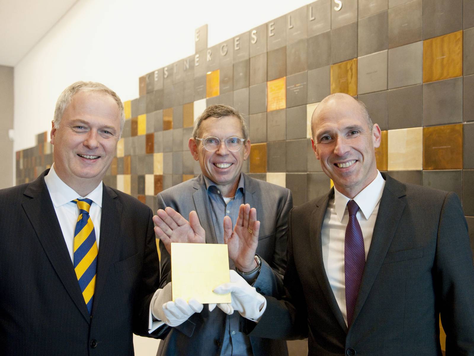 Bankvorstand Dr. David Lohmann (l.) und Nicolas Hoppe (r.) präsentieren gemeinsam mit Veit Görner (Mitte), scheidender Direktor der Kestnergesellschaft, die goldene Kachel.